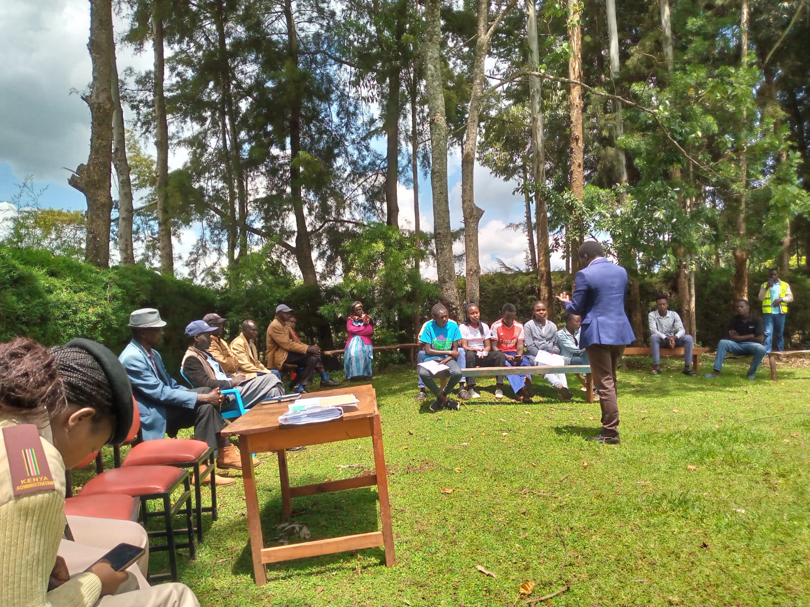 Facilitator speaking to students and guests seated under trees during an outdoor Emgwen TVC information session.