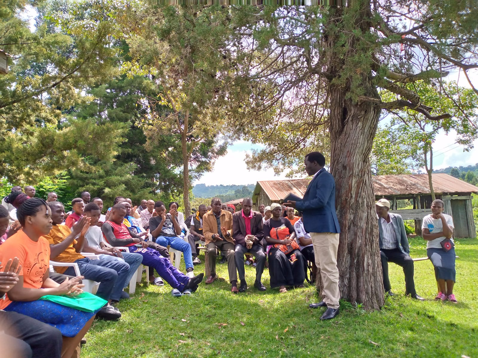 Audience seated outdoors under a tree listening to a speaker during an Emgwen TVC student guidance talk.