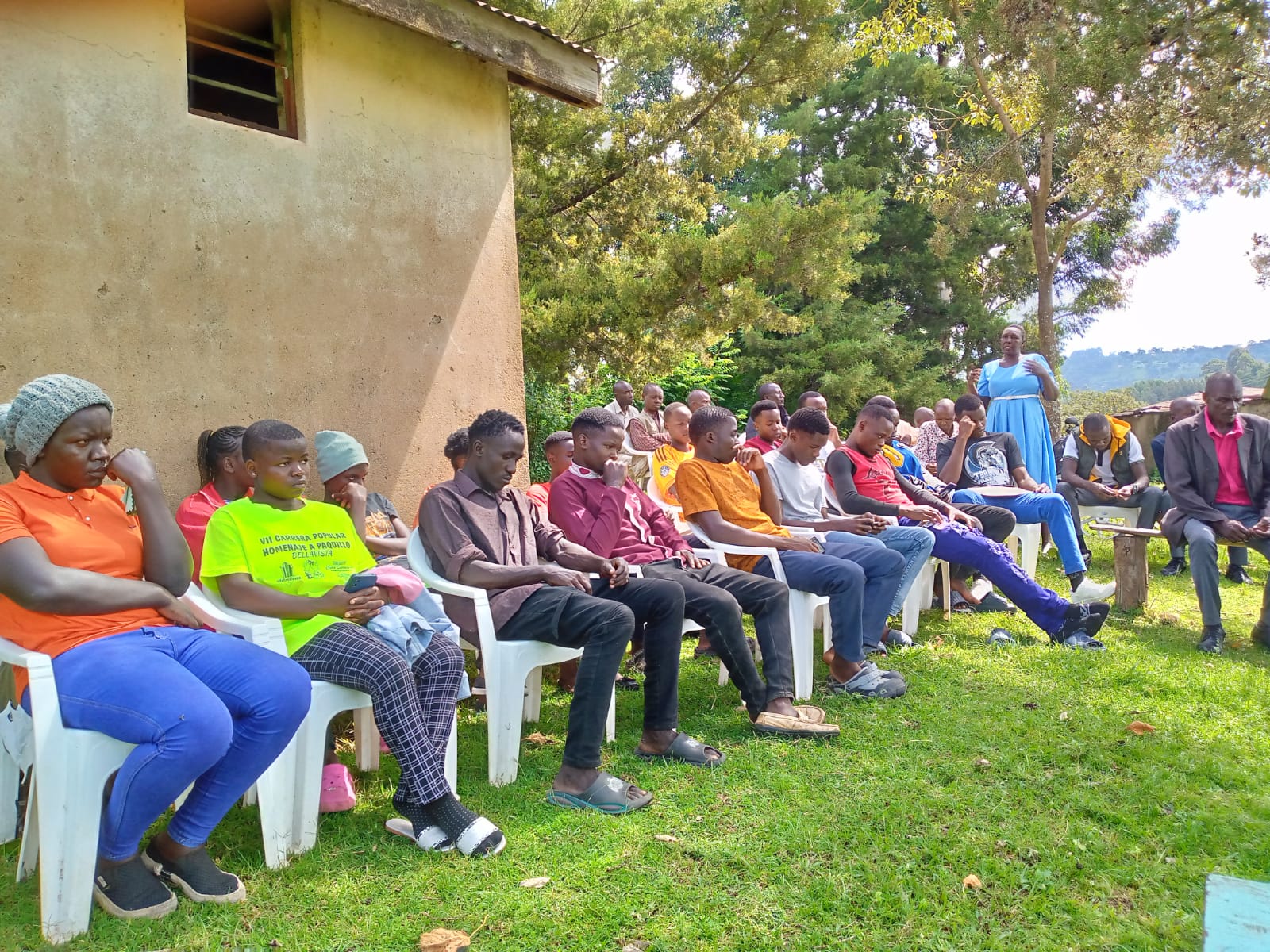 Students and guests seated in a row outdoors near a building while listening during an Emgwen TVC campus session.
