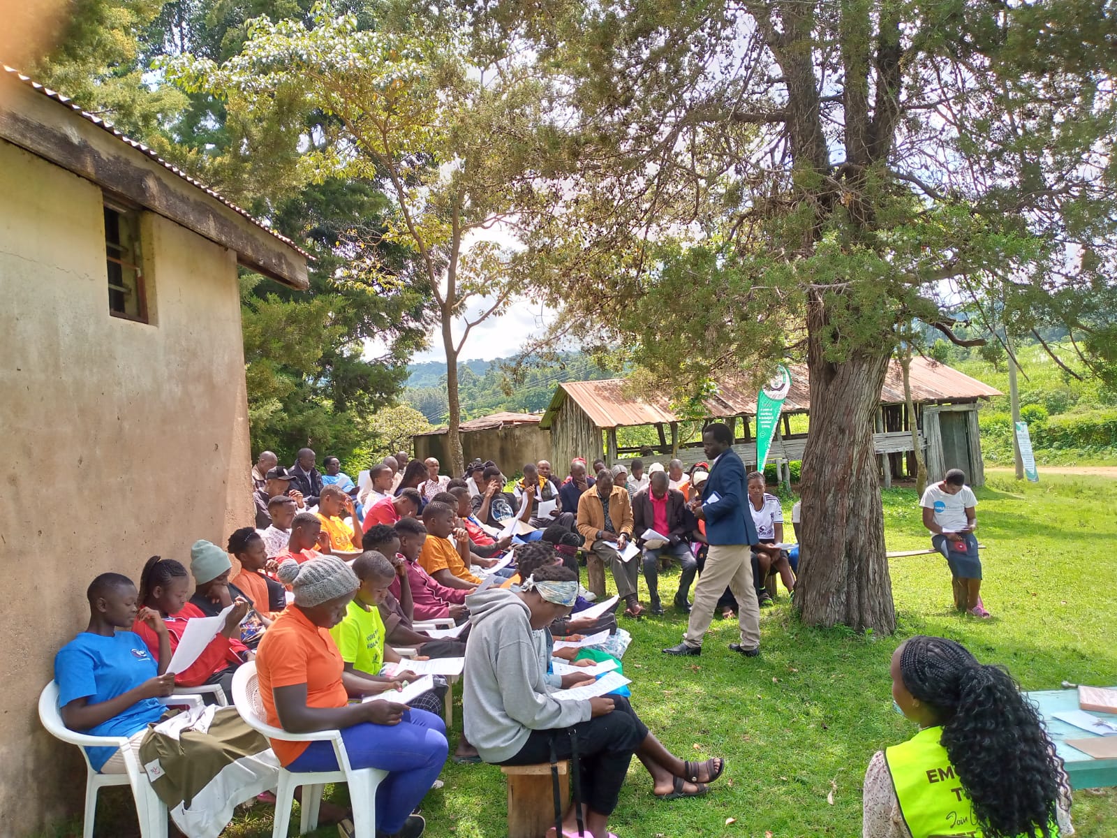 Students seated outside beside a classroom block under trees during an Emgwen TVC student briefing.