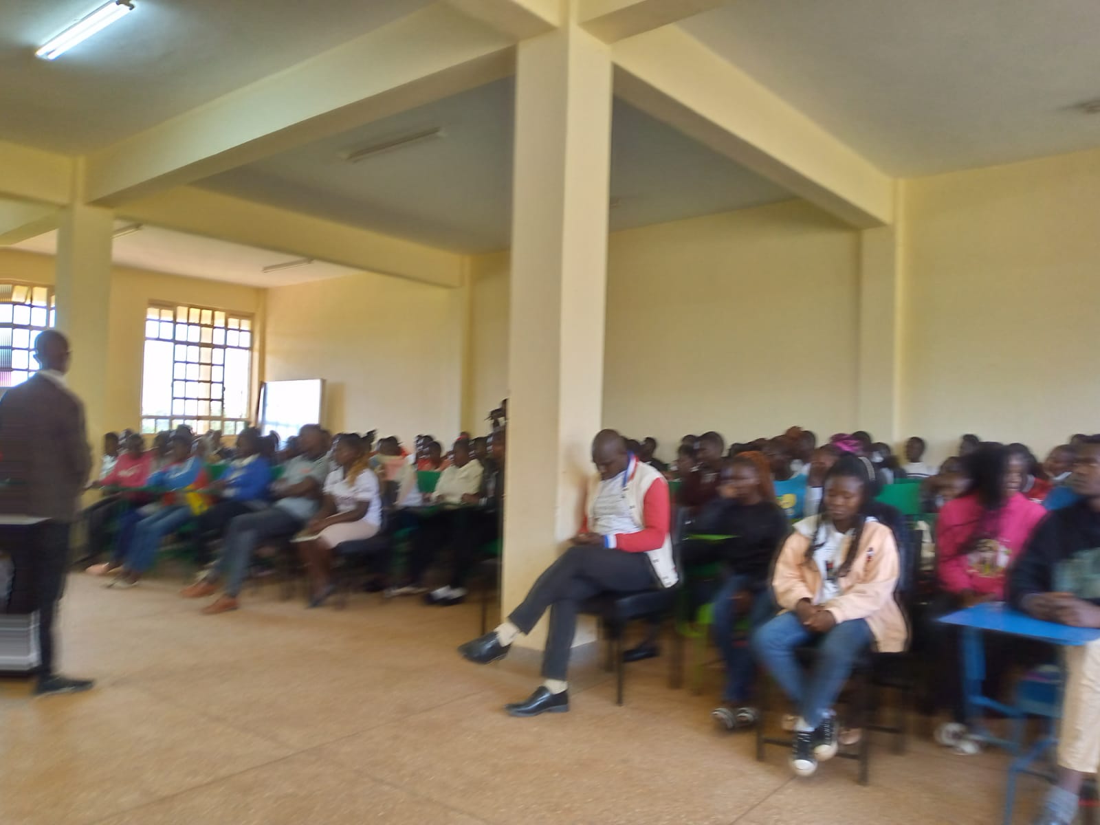 Facilitator standing before students seated inside an Emgwen TVC hall during an orientation discussion.